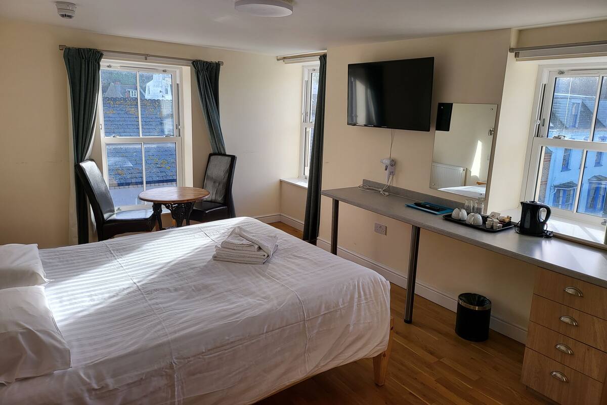 Ein Hotelzimmer mit einem Bett, einem Schreibtisch mit Flachbildfernseher, einem Sessel, einem kleinen Tisch und einem Fenster mit Vorhängen.