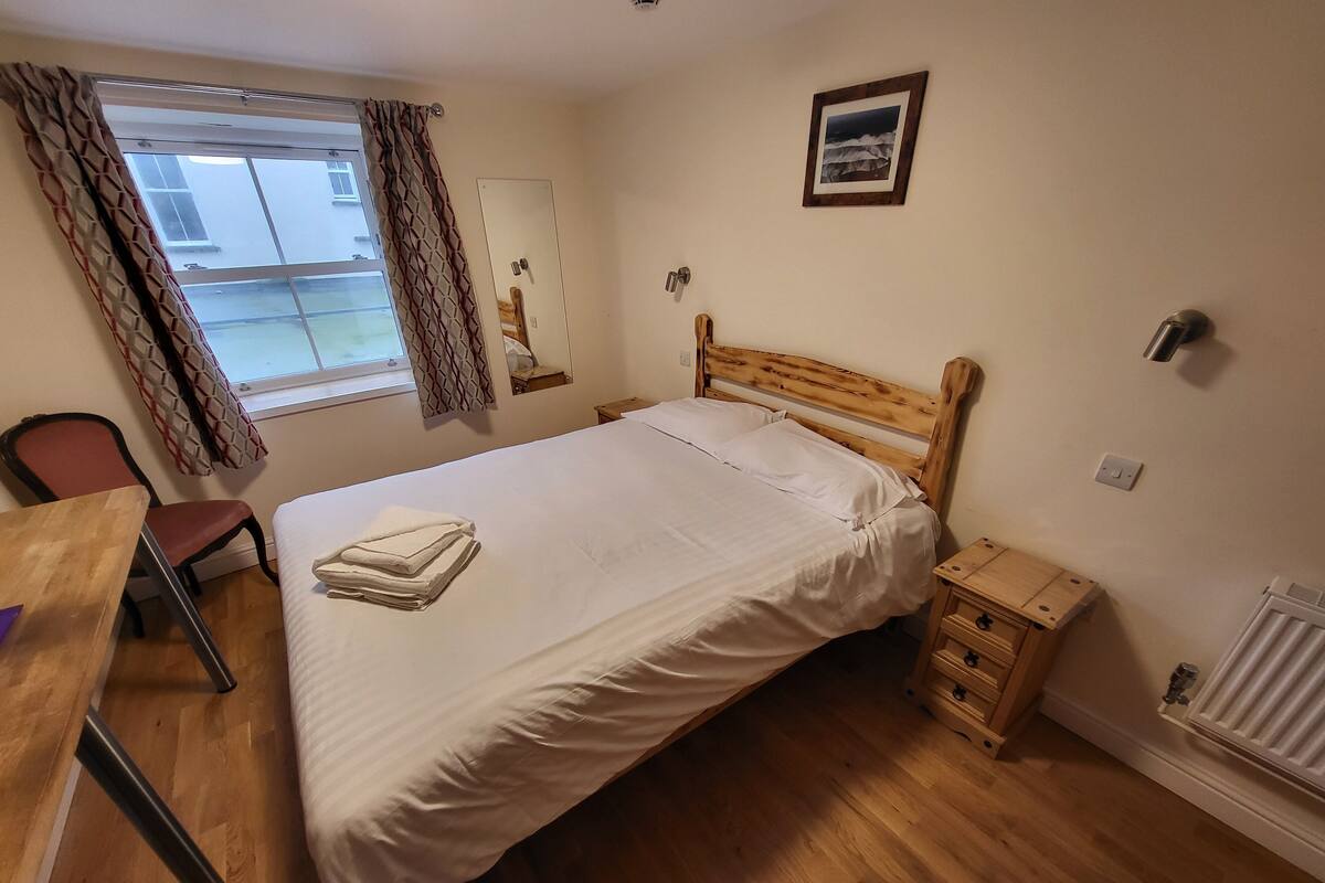 Ein Schlafzimmer mit einem Holzbett, einem kleinen Nachttisch, einem Stuhl, einem Schreibtisch und einem Fenster mit Vorhängen.