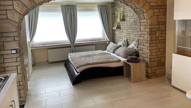 A bedroom with a stone archway, a bed with a headboard, a nightstand, a dresser, and a window with curtains. - 1a apartment Sturm (Emmerthal)