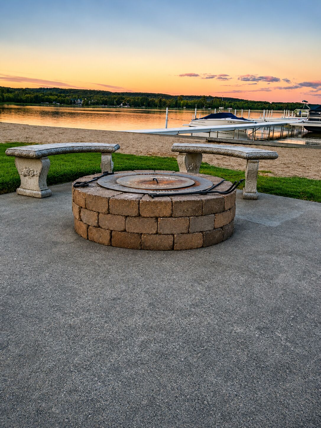Un foyer circulaire en pierre, agrémenté de bancs de chaque côté, installé sur une surface en béton près d’une étendue d’eau.