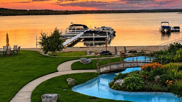 A lakeside view with a wooden dock, a bridge, a swimming pool, and a grassy area with flowers.