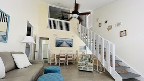 A spacious living area with a high ceiling, a ceiling fan, a dining table with chairs, a staircase with white railings, and a large window with blinds.