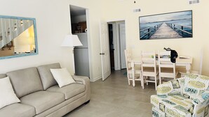 A living room with a beige sofa, a dining table with chairs, a mirror, and a painting of a pier.