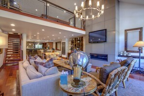 Relax in this elegantly furnished living area with a high ceiling and mezzanine level, featuring a flat-screen TV, plush sofa, and wicker armchair.