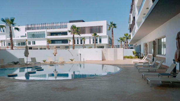 A modern hotel pool area with sun loungers and umbrellas, surrounded by palm trees and a multi-story building with large windows. - Zephyr Agadir (Agadir)