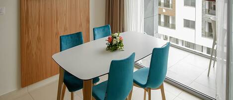 Un comedor moderno con una mesa blanca, cuatro sillas azules y un centro de mesa floral. Un televisor de pantalla plana está montado en la pared, y la habitación tiene ventanas amplias con vistas a un edificio.