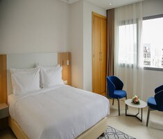 Een hotelkamer met een groot bed, twee blauwe stoelen, een klein rond tafeltje met een bloem, en een houten garderobe.