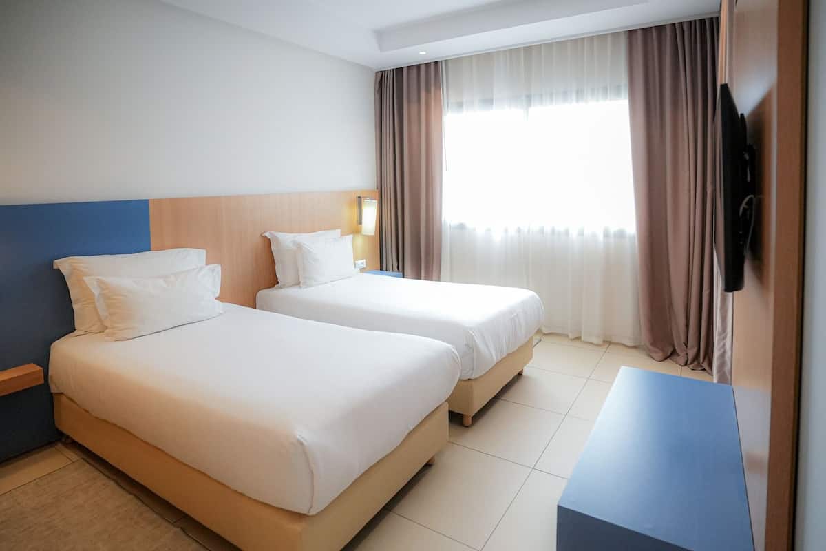 Ett hotellrum med två sängar, en platt-TV och ett stort fönster med gardiner.