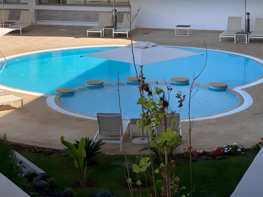 Una piscina con sillas de descanso y sombrillas, rodeada de un jardín y un edificio.