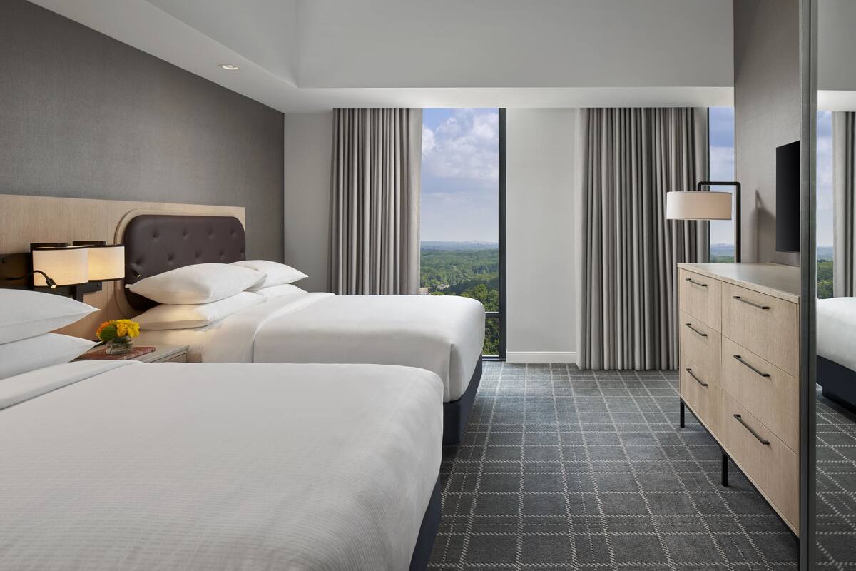 Quarto de hotel com duas camas, cômoda, televisão e uma janela grande com cortinas.