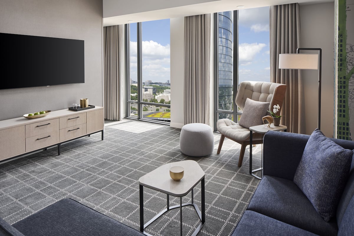 Quarto de hotel moderno com uma janela grande, televisão de tela plana, sofá, poltrona, mesa pequena e mesa de cabeceira.