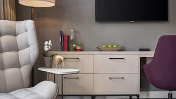 A modern living room with a grey sofa, a small round table, a desk with drawers, a purple chair, a flat-screen TV mounted on the wall, and a bowl of apples on the desk.
