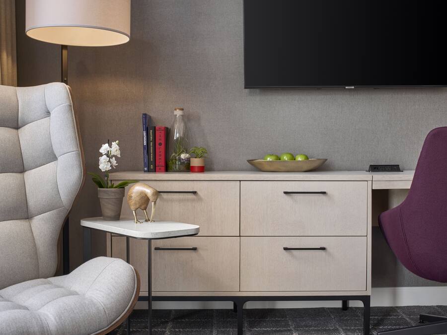 A modern living room with a grey sofa, a small round table, a desk with drawers, a purple chair, a flat-screen TV mounted on the wall, and a bowl of apples on the desk.