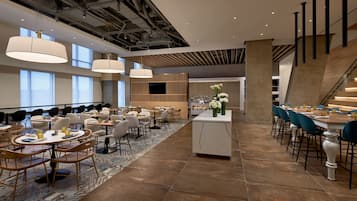 A modern hotel lobby with round tables, chairs, and hanging lamps. There is a staircase on the right and a flat-screen TV on the wall.