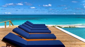 A wooden deck with blue lounge chairs facing a clear blue ocean and a swimming pool. - BeauSoleil Beach House, Luxury Private Ocean Front with Infinity Pool/Hot Tub (Exuma)
