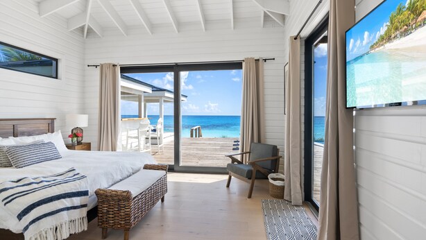 A bedroom with a large bed, a chair, a small table, and a view of the beach through a sliding door. - BeauSoleil Beach House, Luxury Private Ocean Front with Infinity Pool/Hot Tub (Exuma)