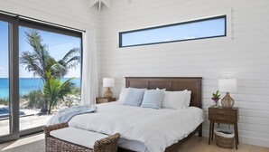 A bedroom with a large window overlooking a beach, a bed with white linens, a wicker chair, and a nightstand with a lamp. - BeauSoleil Beach House, Luxury Private Ocean Front with Infinity Pool/Hot Tub (Exuma)