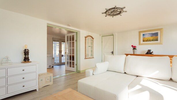 A spacious living room with a white sofa, wooden coffee table, and a white dresser. A door leads to another room, and a framed picture hangs on the wall. - Plum Guide - New Tricks (Norwich)