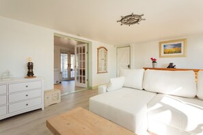 A spacious living room with a white sofa, wooden coffee table, and a white dresser. A door leads to another room, and a framed picture hangs on the wall. - Plum Guide - New Tricks (Norwich)