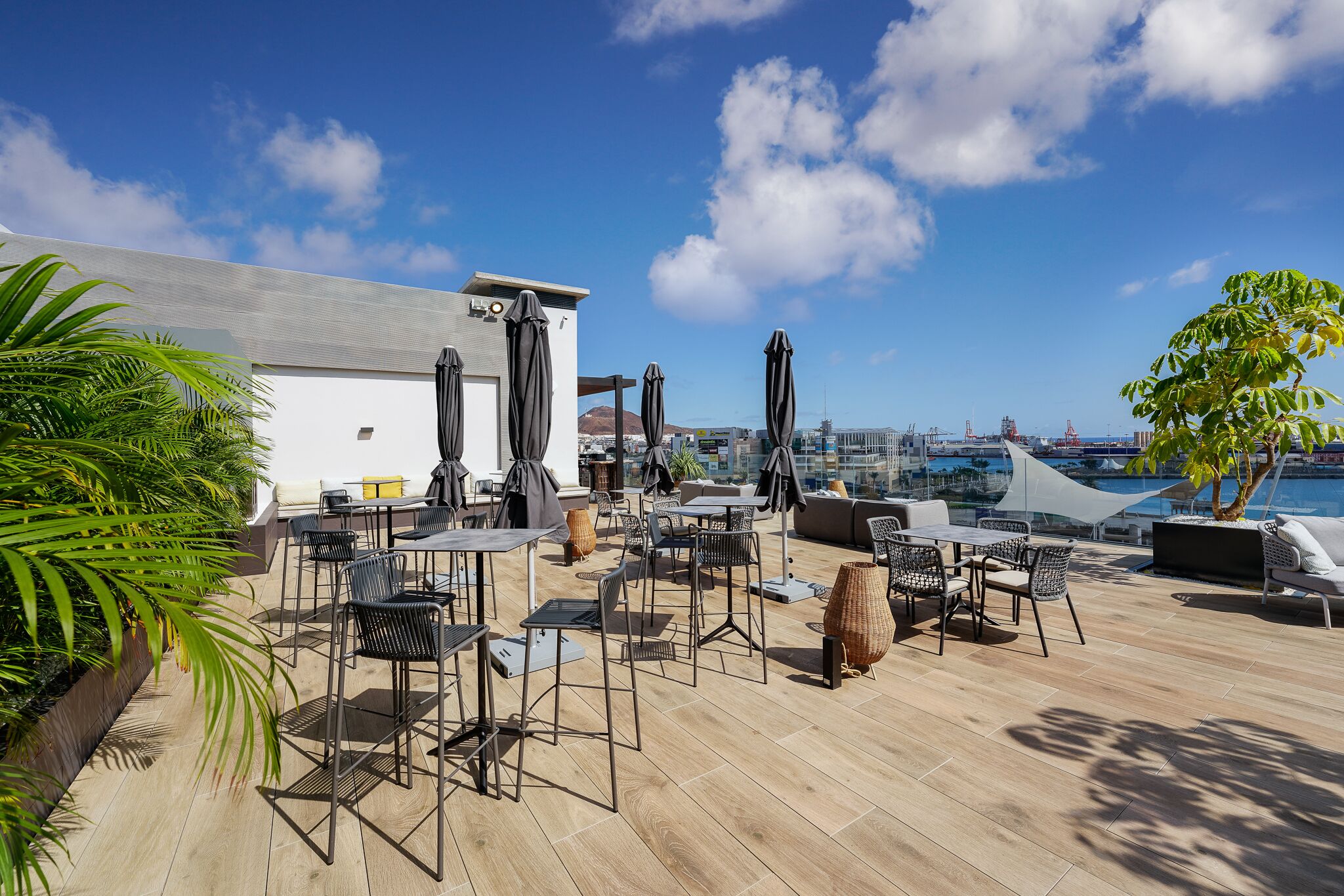 An outdoor terrace with tables and chairs, a white building, and a view of the city and water.