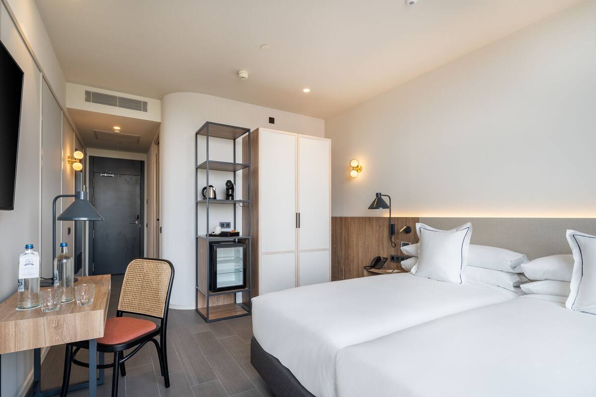 A modern hotel room with a bed, a desk with a chair, a wardrobe, and a small refrigerator.