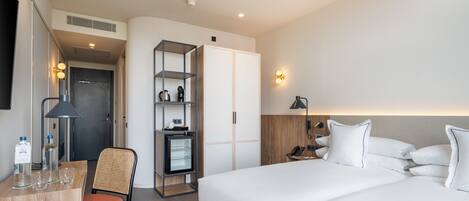 A modern hotel room with a bed, a desk with a chair, a wardrobe, and a small refrigerator.