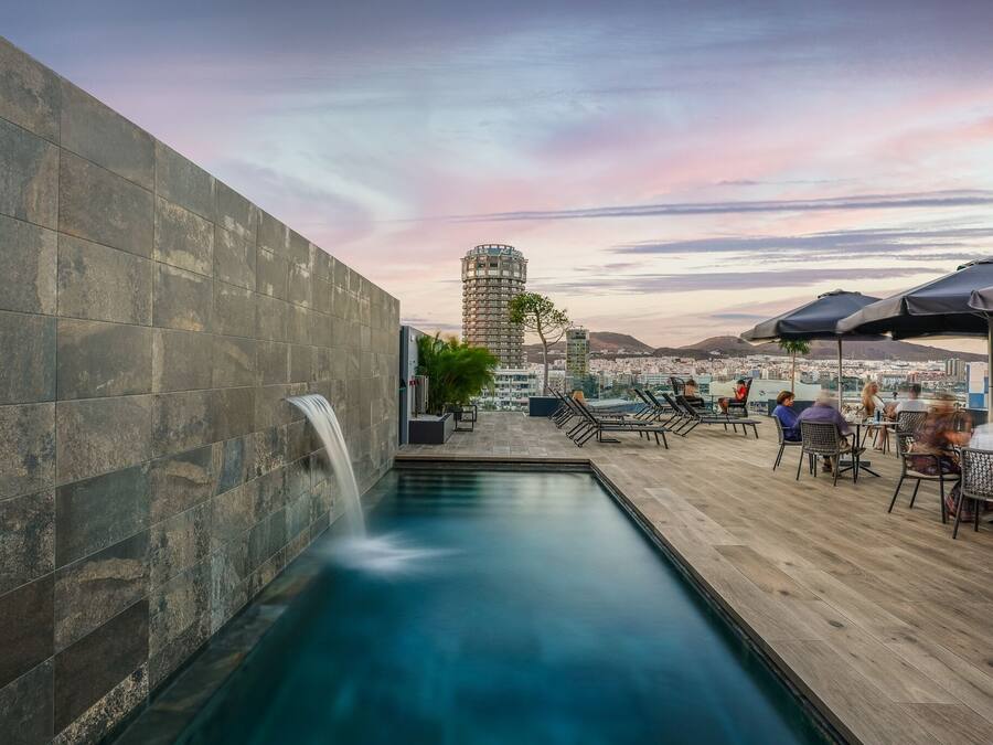 A rooftop pool with a waterfall, outdoor seating, and a view of the cityscape.