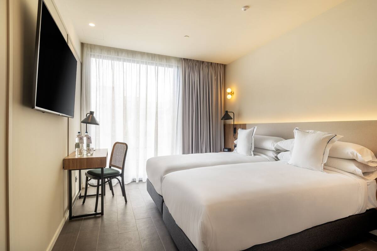 A hotel room with two beds, a desk with a chair, a flat-screen TV, and a large window with curtains.