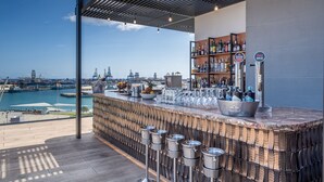 A rooftop bar with a city skyline view, bar stools, and a variety of bottles and glasses.