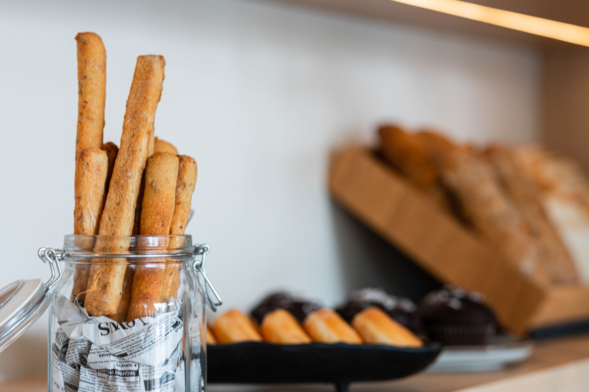 A glass jar filled with baguettes, a tray of pastries, and a loaf of bread in the background.