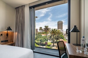 Una habitación de hotel con un ventanal grande, una cama, un escritorio con silla y una lámpara.