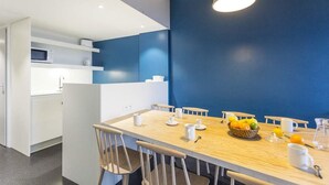 Zona de menjador moderna amb taula i cadires de fusta, cistella de fruita i un espai per al cafè amb elements blancs.