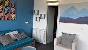 Habitació amb un sofà blau, una butaca blanca, un radiador i una porta que dona a una altra estança.