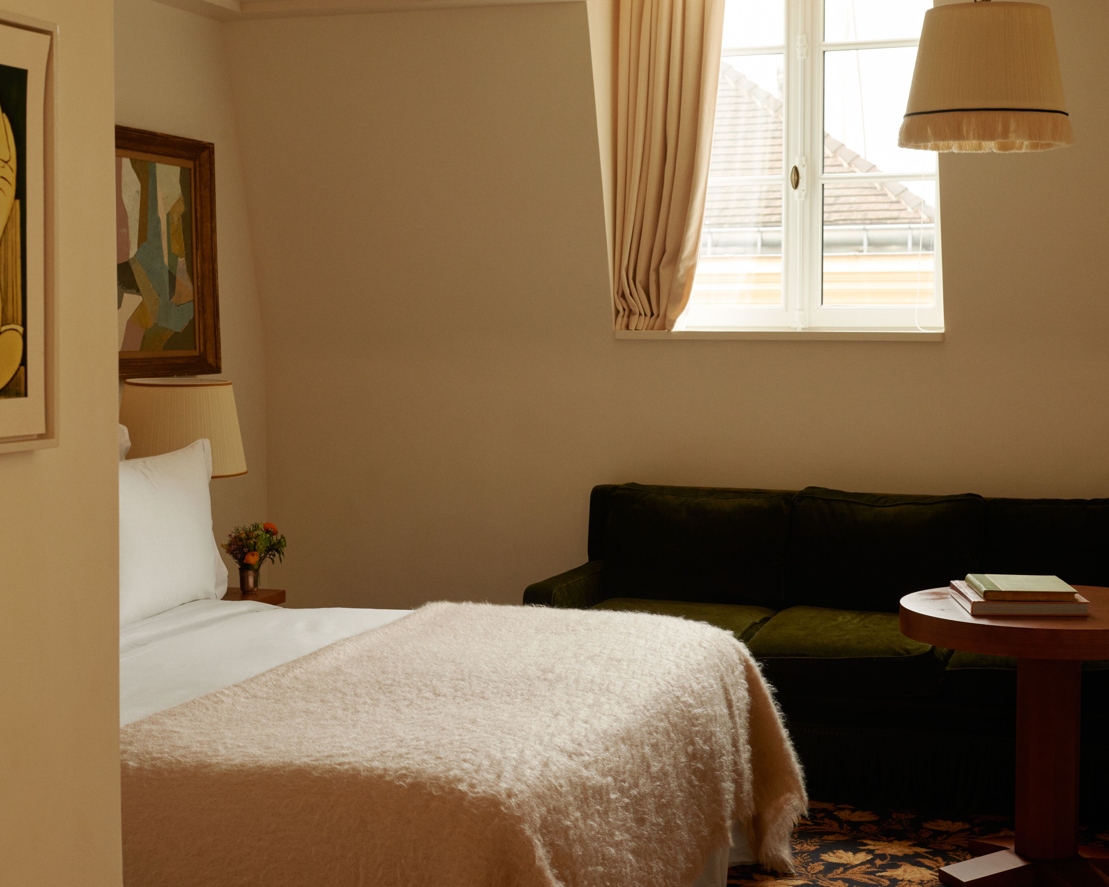 Una habitación de hotel con una cama, un sofá verde, una mesita con libros, una lámpara y un cuadro en la pared.