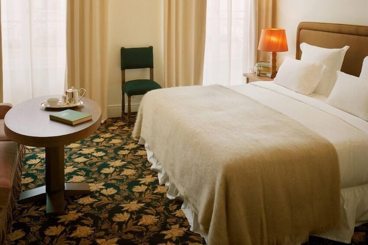 A hotel room with a bed, a round table with a book and teapot, a chair, and a lamp.