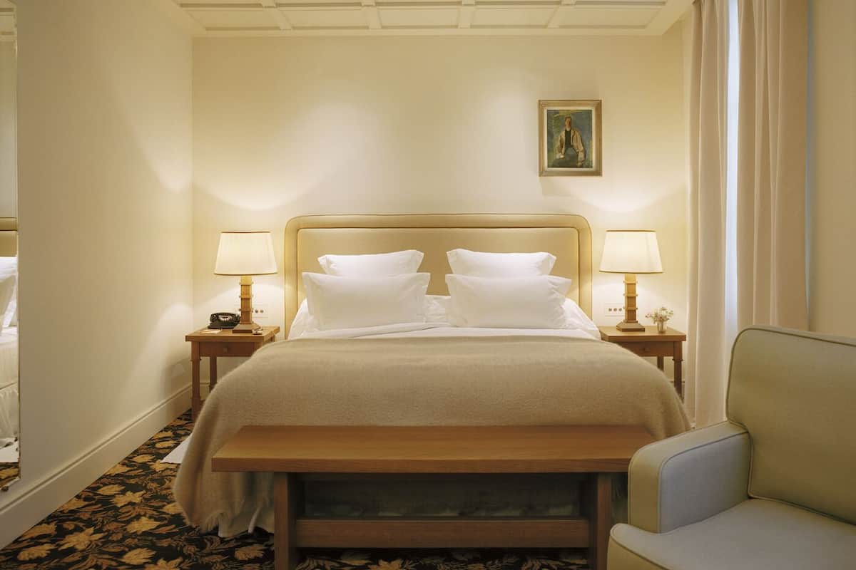 A hotel room with a large bed, two bedside tables with lamps, a bench, a chair, and a painting on the wall.