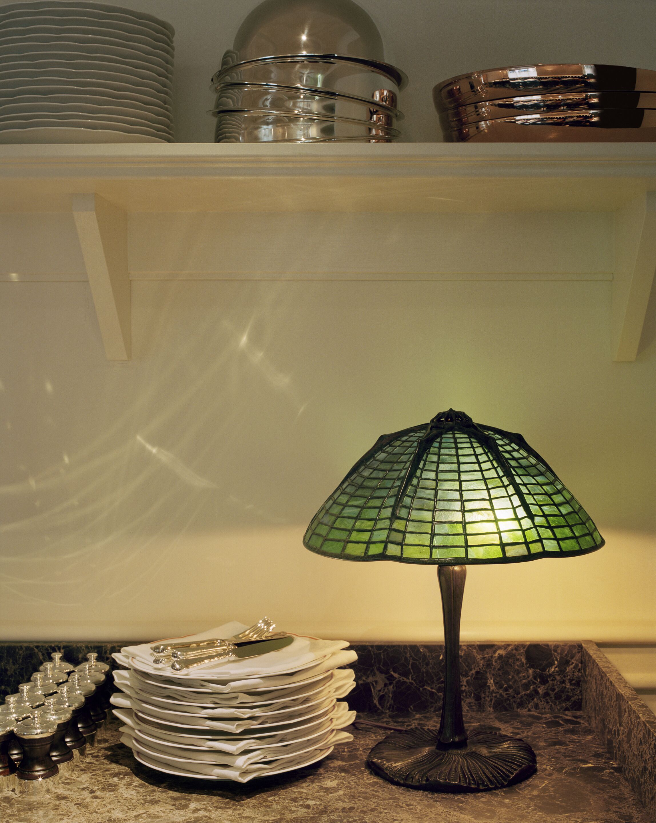 Un piano di lavoro in cucina con una lampada a mosaico verde, piatti impilati e una ciotola decorativa.