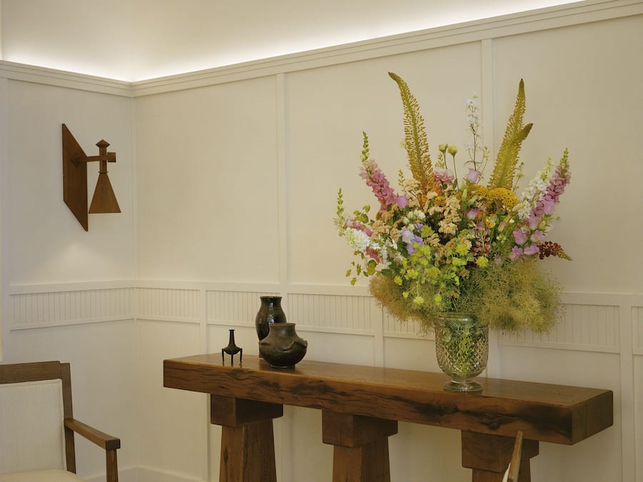 A room with a wooden table, two chairs, a vase with flowers, and a wall-mounted sculpture.