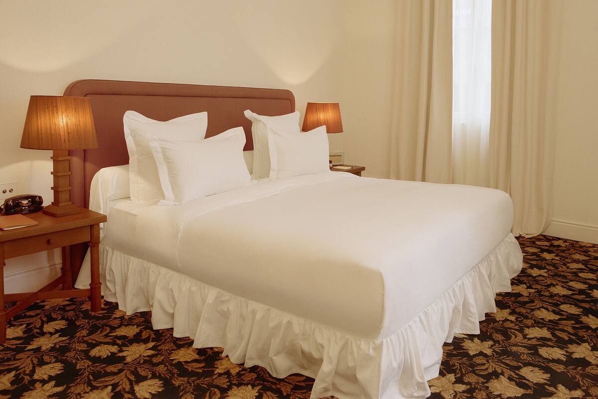 A hotel room with a large bed, two bedside tables, a nightstand, a lamp, and a window with curtains.