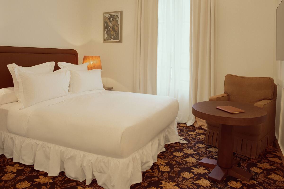A hotel room with a large bed, a bedside table with a lamp, a chair, and a small table with a book.