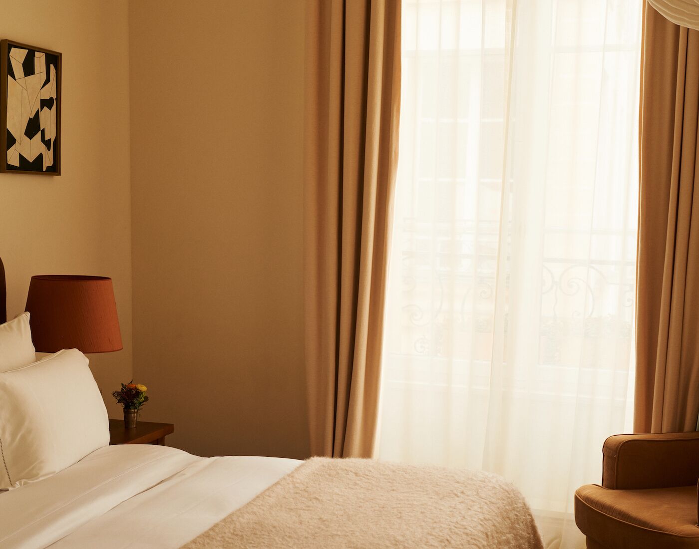 Een hotelkamer met een bed, nachtkastje met lamp, stoel en raam met gordijnen.