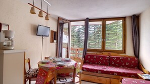 A cozy room with a wooden table set for dining, a red patterned sofa, a television mounted on the wall, and large windows with curtains. - travelski home classic - Résidence Boedette - Studio 4 personnes 515 (Les Menuires)