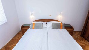 Ein Doppelbett mit weißen Leinen und zwei Kissen, an beiden Seiten Nachttische mit Lampen, in einem Zimmer mit Holzboden und schlichten Wänden.