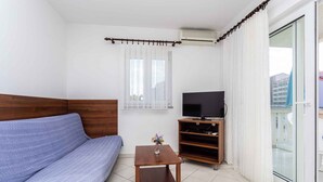 Ein kleiner, moderner Raum mit einer Couch, einem Fernseher auf einem Schrank, einem kleinen Tisch mit einer Vase und einer Glastür zum Außenbereich.