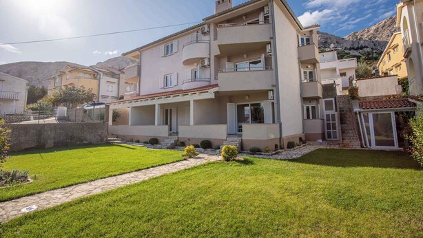 Um edifício residencial de vários andares com varandas, um pátio pavimentado e montanhas ao fundo.