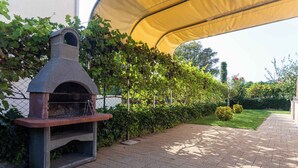 A stone outdoor pizza oven under a yellow canopy, surrounded by a hedge and a paved walkway. - Holiday apartment only 100 m from the sea (Baška)