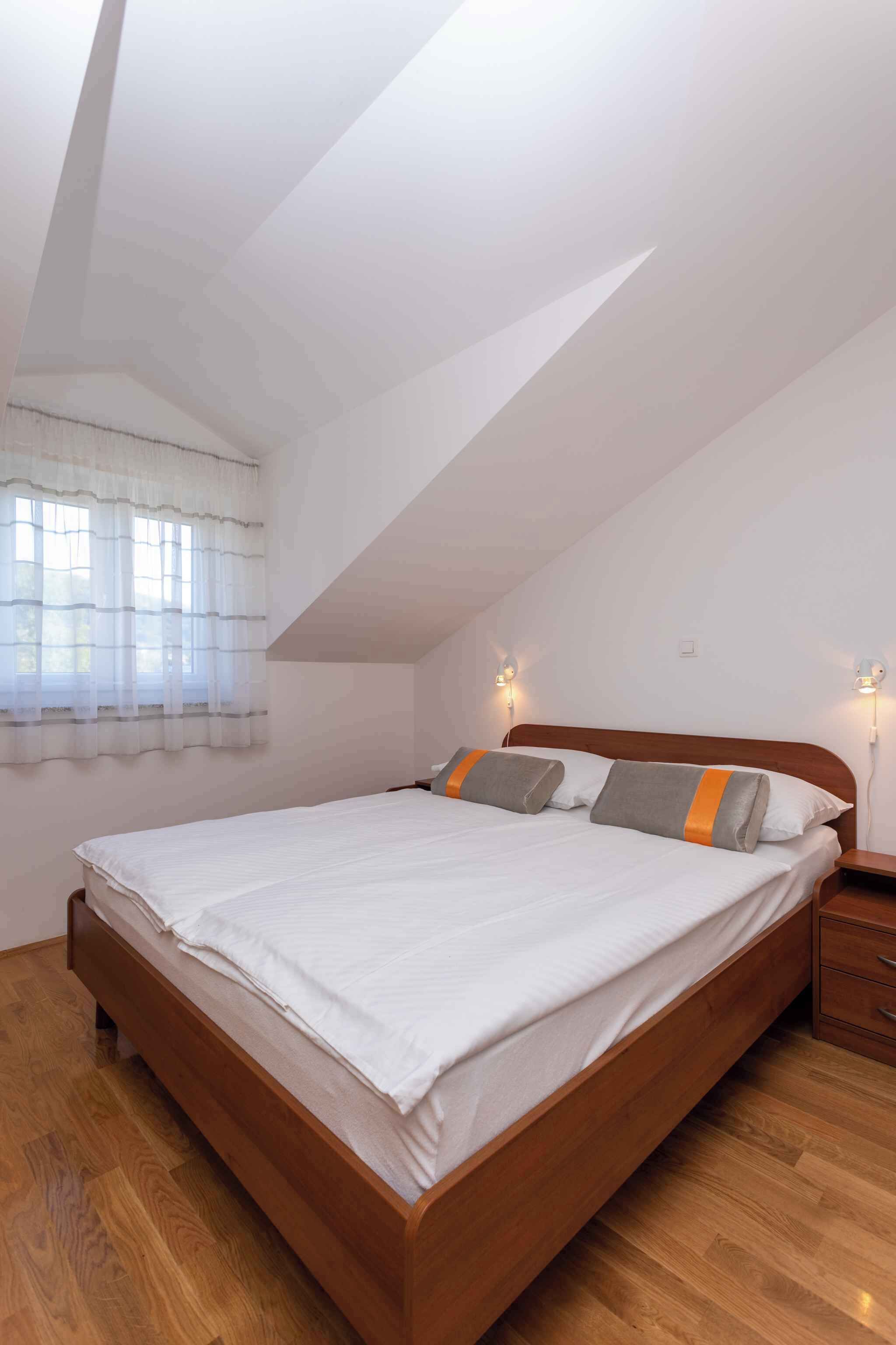 Un dormitorio con cama de madera, ropa de cama blanca, una ventana con cortinas traslúcidas y luces fijadas a la pared.