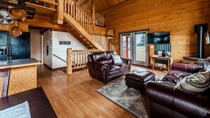 A wooden cabin interior with a staircase, leather sofas, a kitchen area, and a fireplace. - Railroad Baron (Superior)