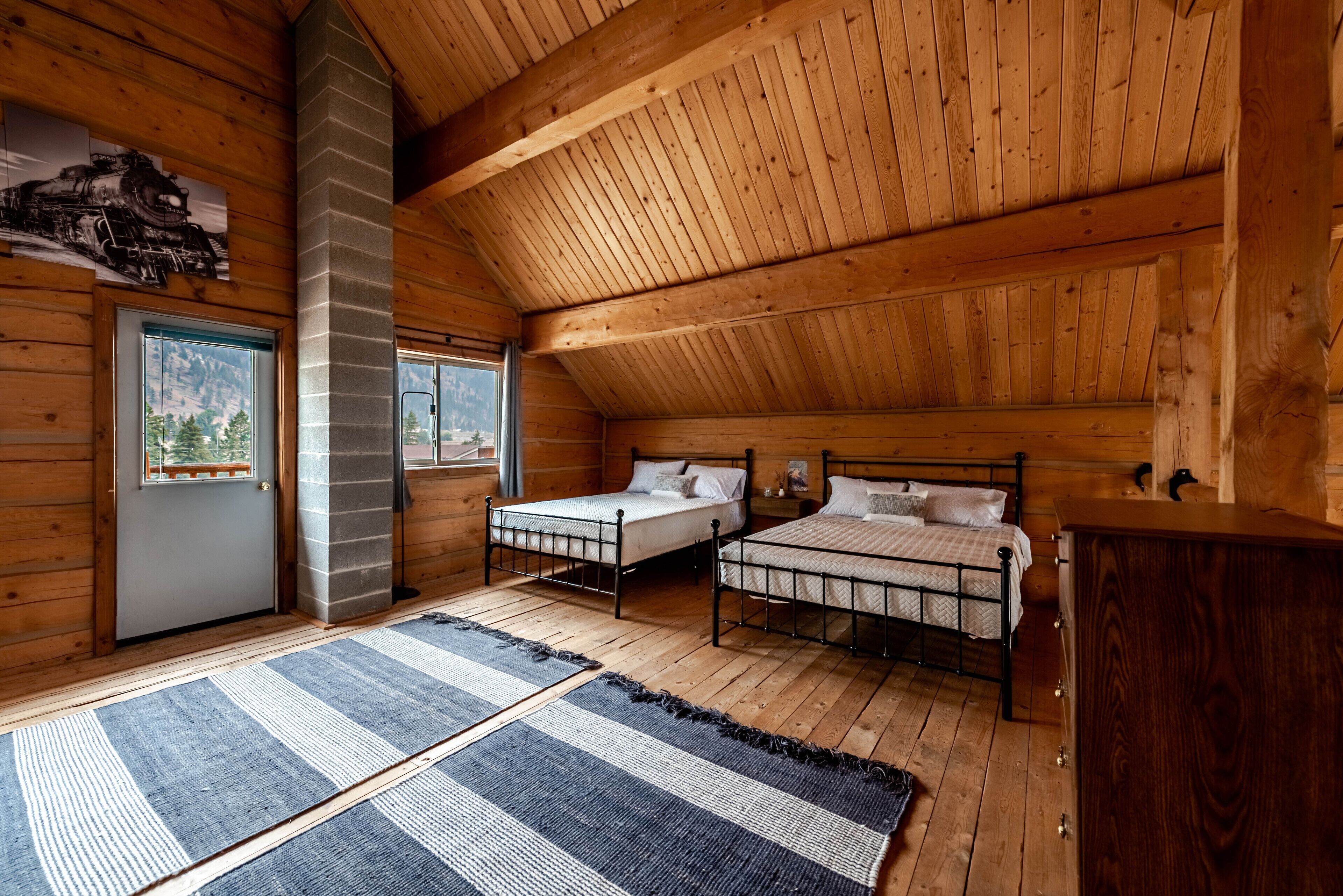 A log cabin bedroom with two metal beds, a fireplace, and a window with a mountain view.