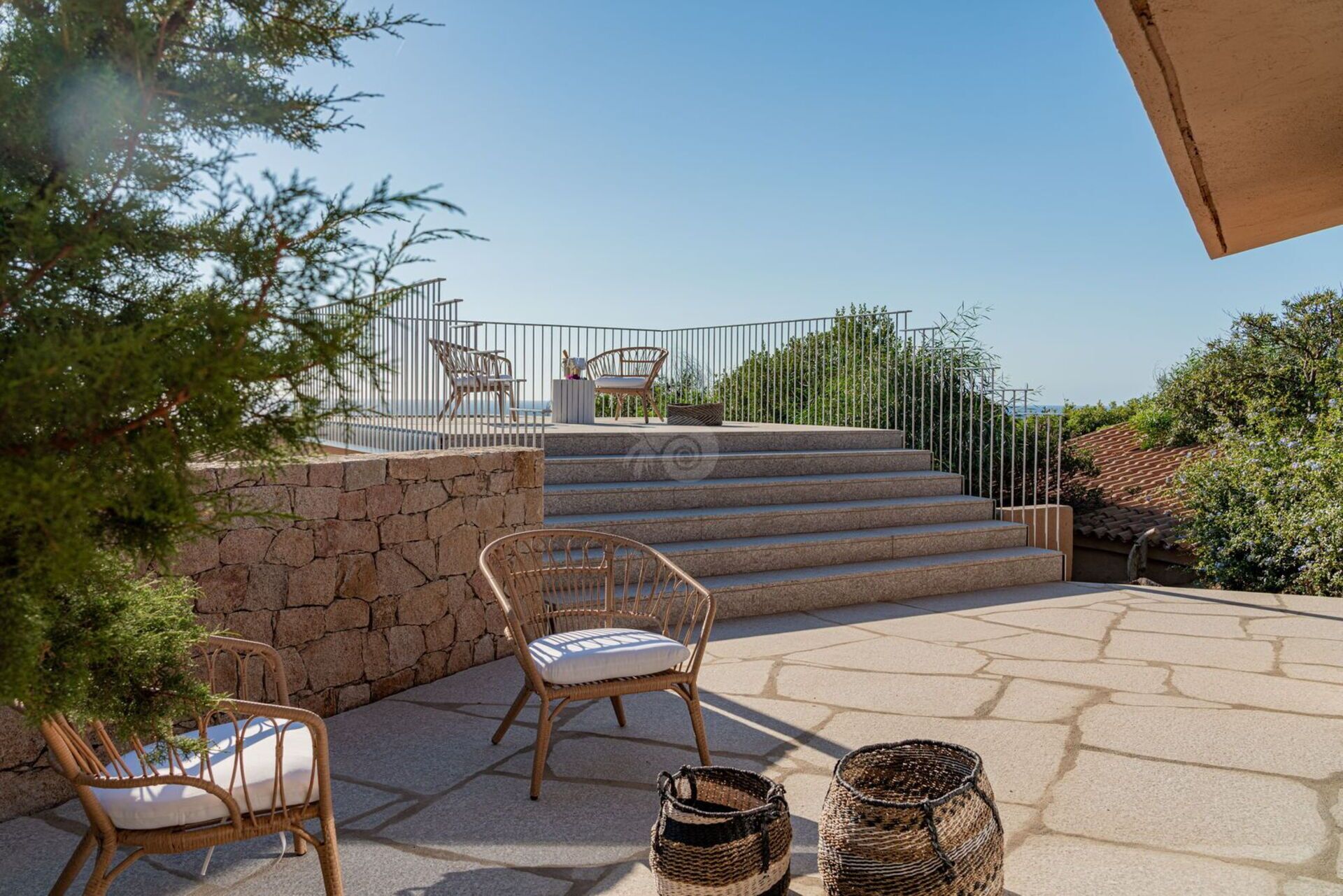 A patio with wicker chairs, a stone wall, and a view of the sea.